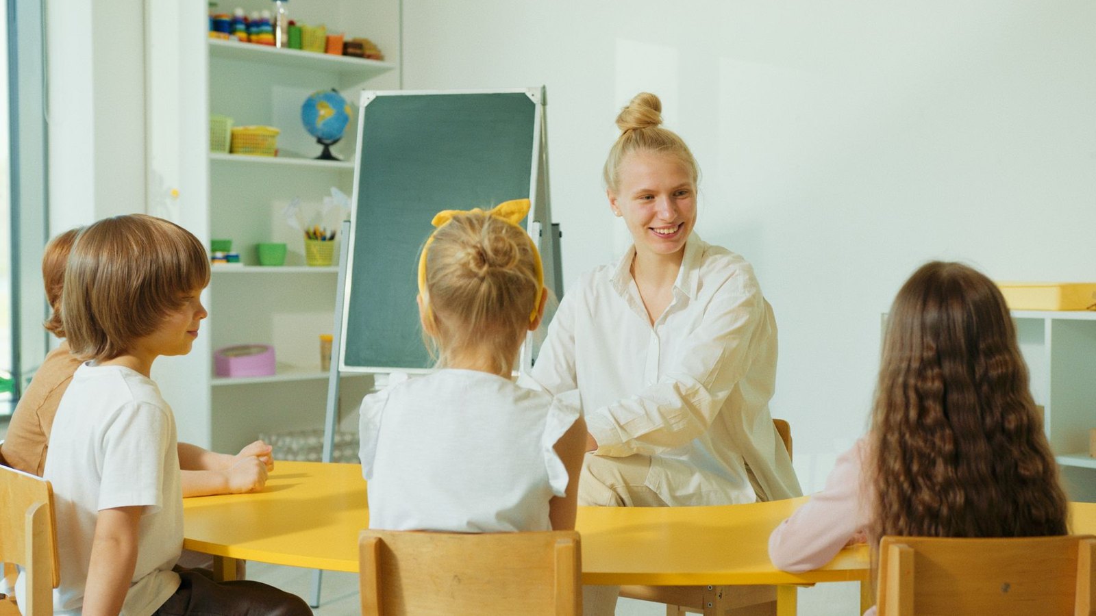 Erzieherin sitzt mit vier Kindern an einem Tisch in der Kita
